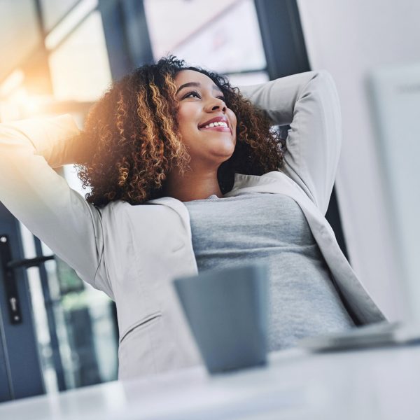 Business women leans back and smiles with arms above head relaxing at her desk