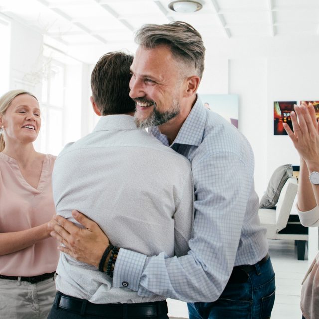 A group cheering and clapping while two men hug in an office