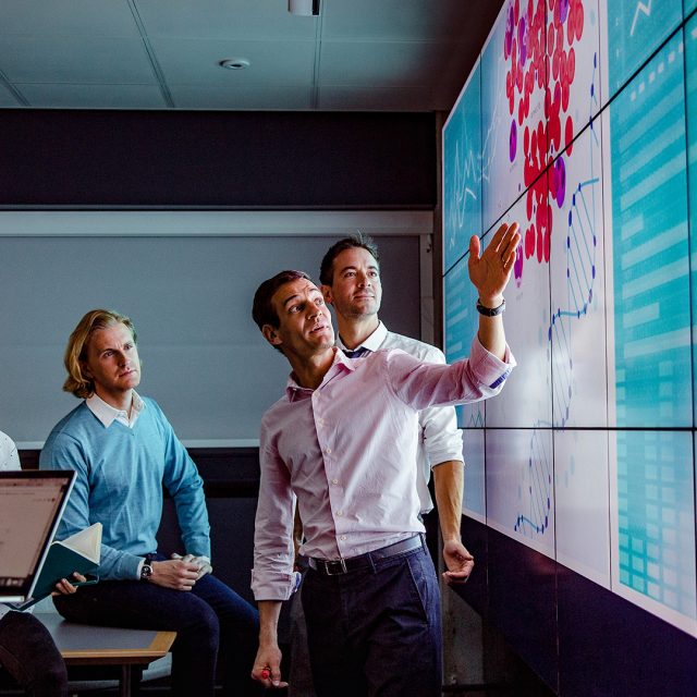 Three men in a presentation, with one man pointing to a colorful graph while speaking and the two others listening